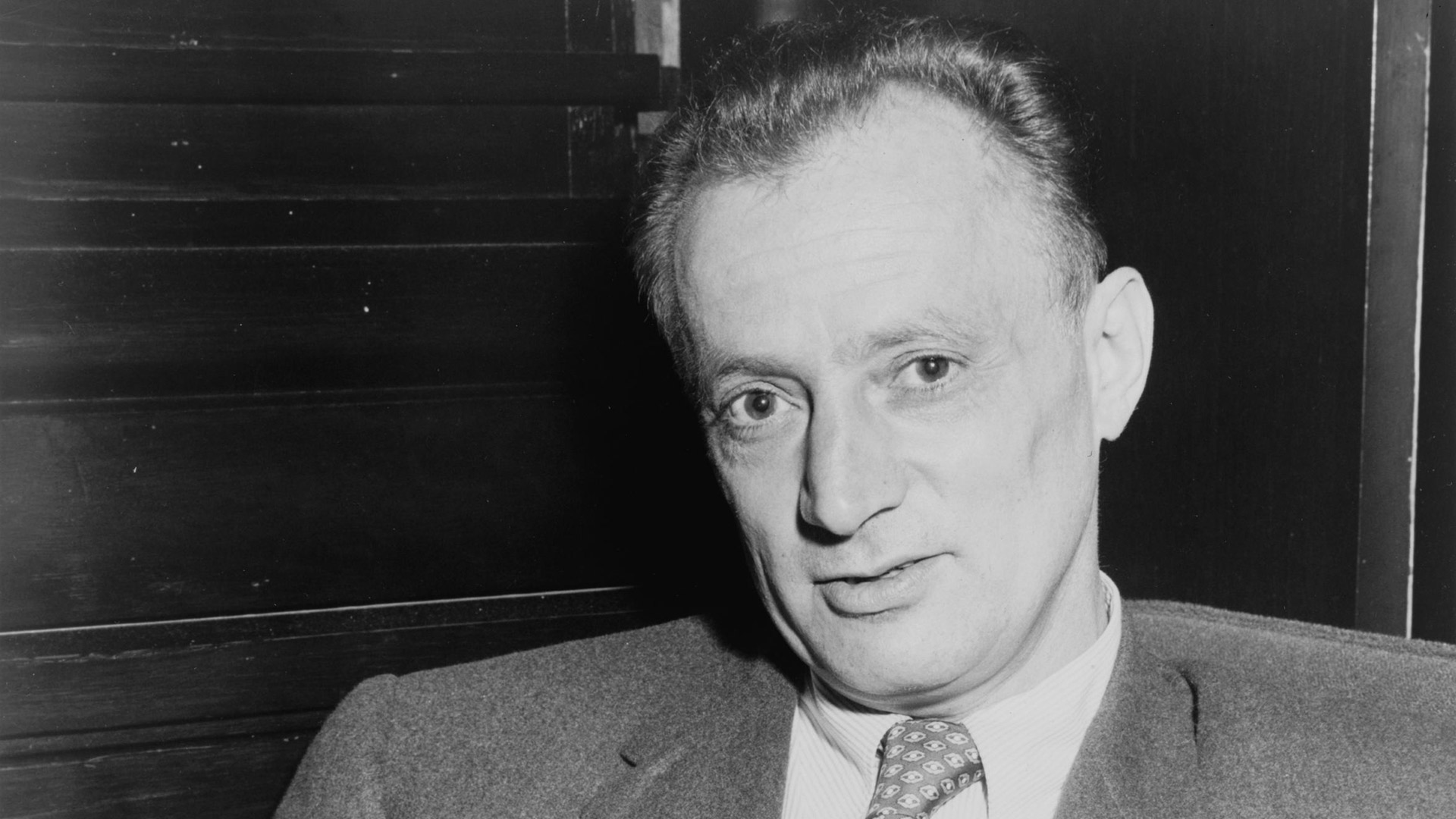 Nelson Algren sitting in a suit looking at the camera and holding a paper