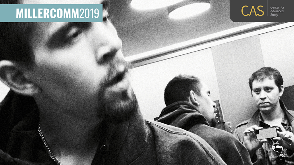 candid black and white photo of two men in a small space; one films the other two on his phone through a mirror