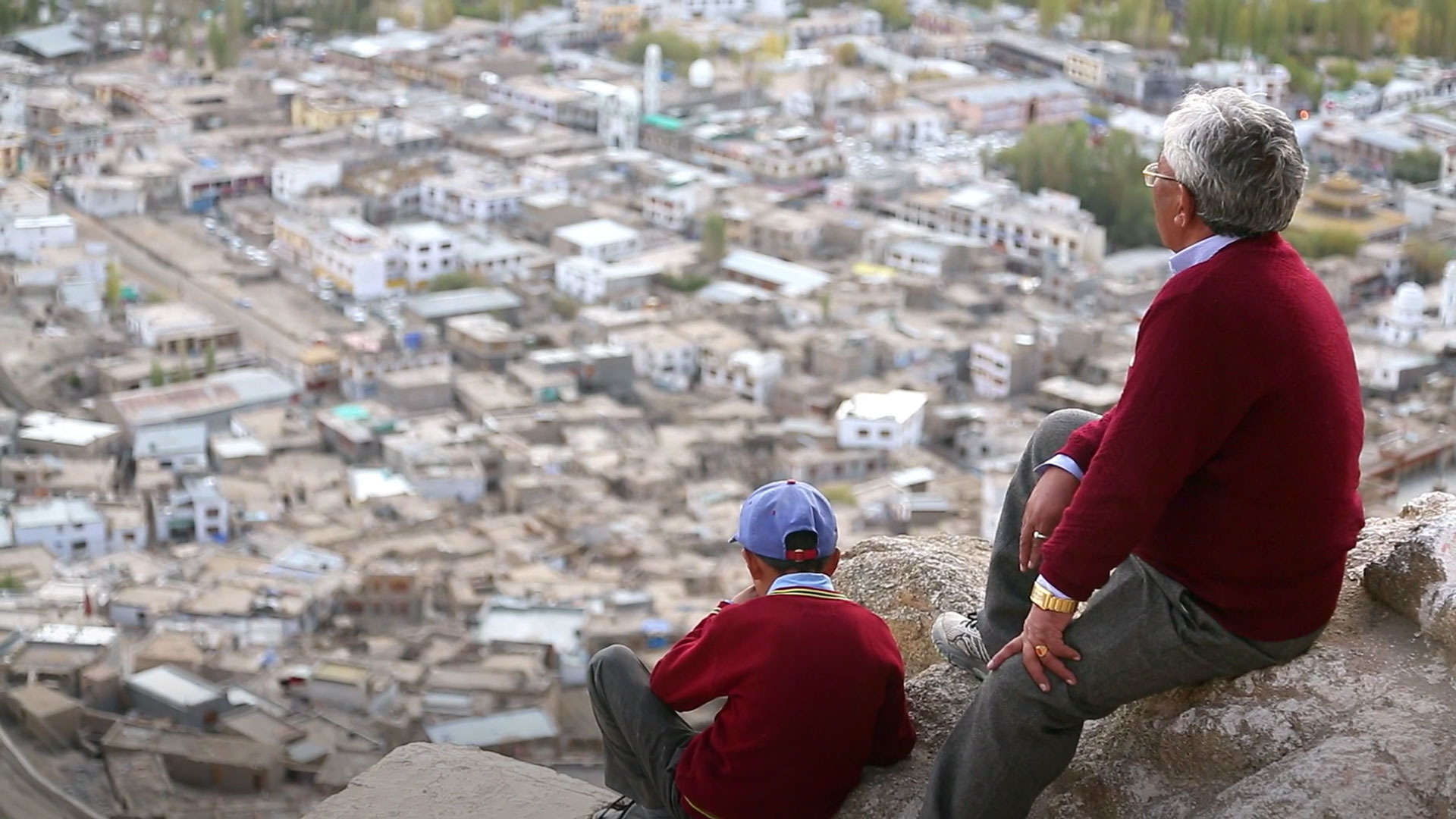 a man and his grandson sit on a rockface overlooking a large, flat city