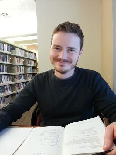 A man smiles at the camera while seated with a book in a library