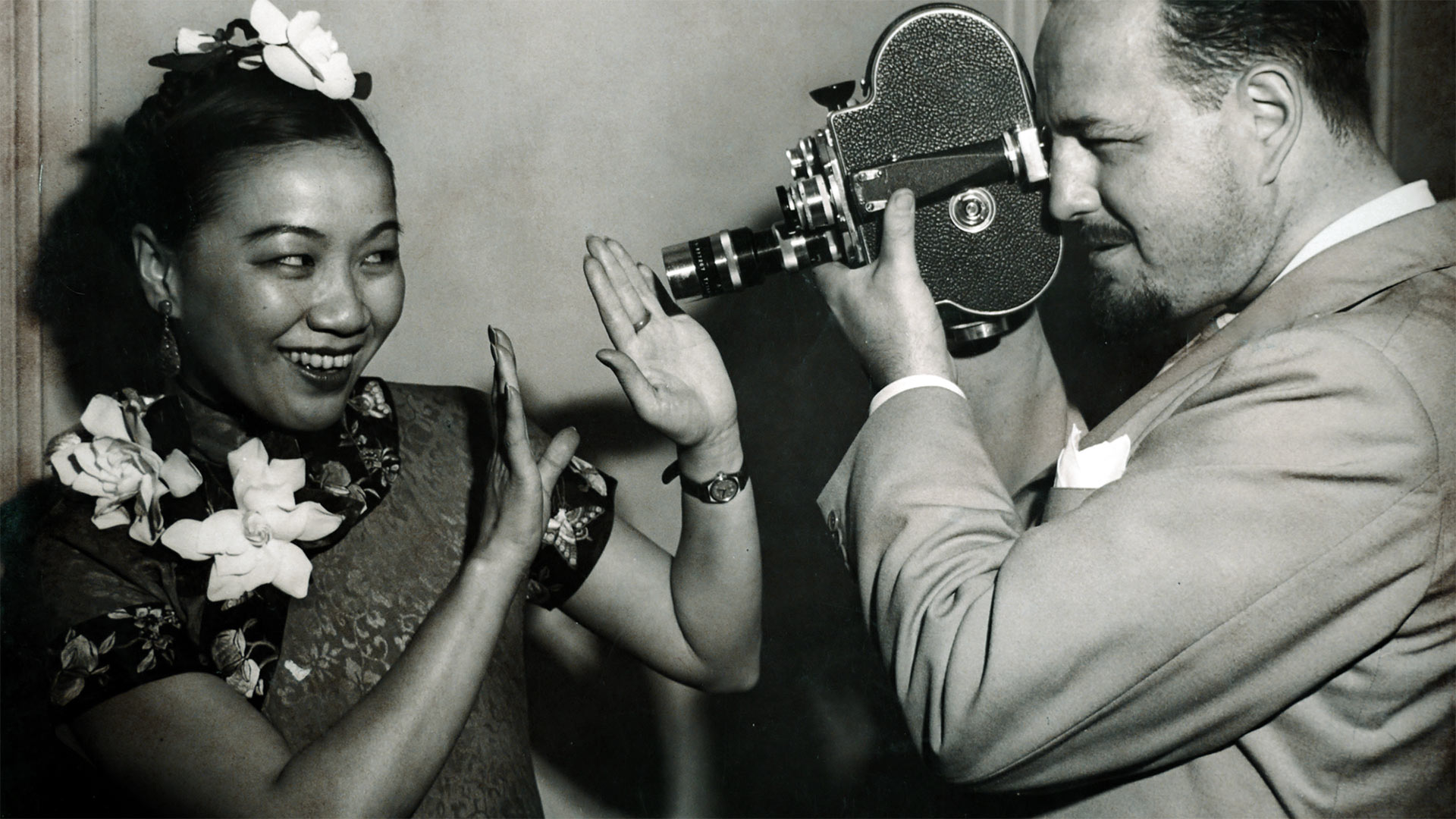 1940s photo of a man with handheld video camera pointing at smiling woman with flowers