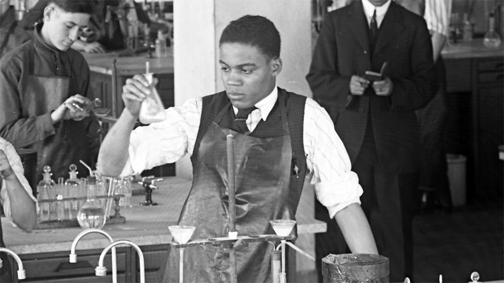 A young man inspects chemistry equipment in an early 20th century chemistry lab