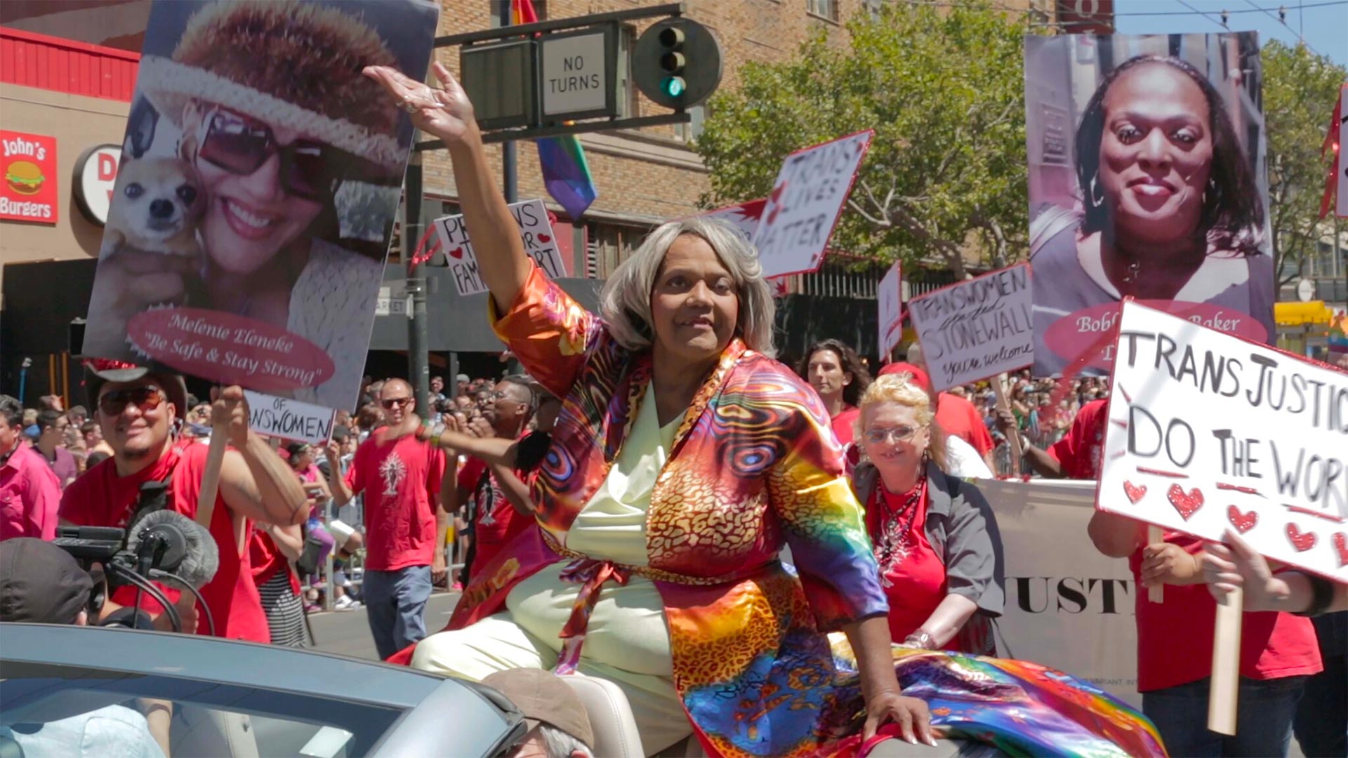 Woman in colorful clothing in a trans-woman march; many people march with smiles and signs