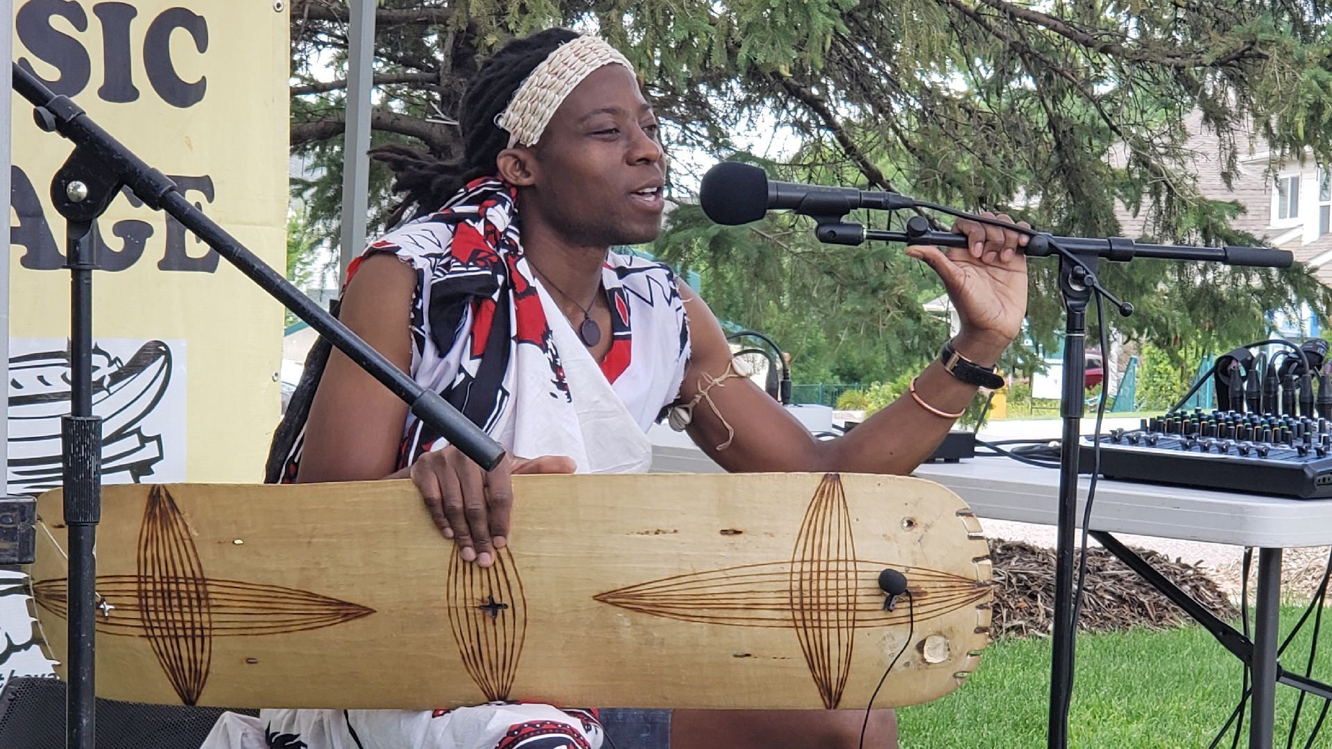 Photo of musician performing in an outside venue, sitting in front of a mic with a large wooden instrument