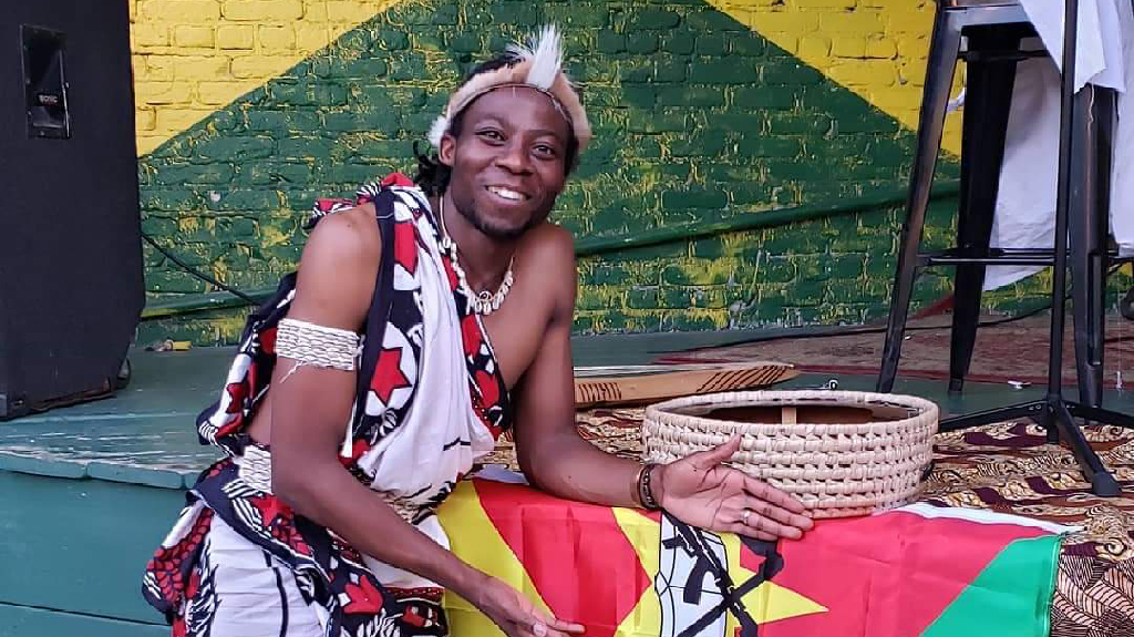 Man dressed in traditional African clothing standing next to colorful flag in front of stage 
