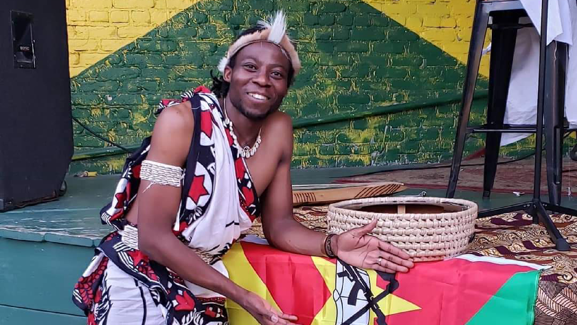 Man dressed in traditional African clothing standing next to colorful flag in front of stage 