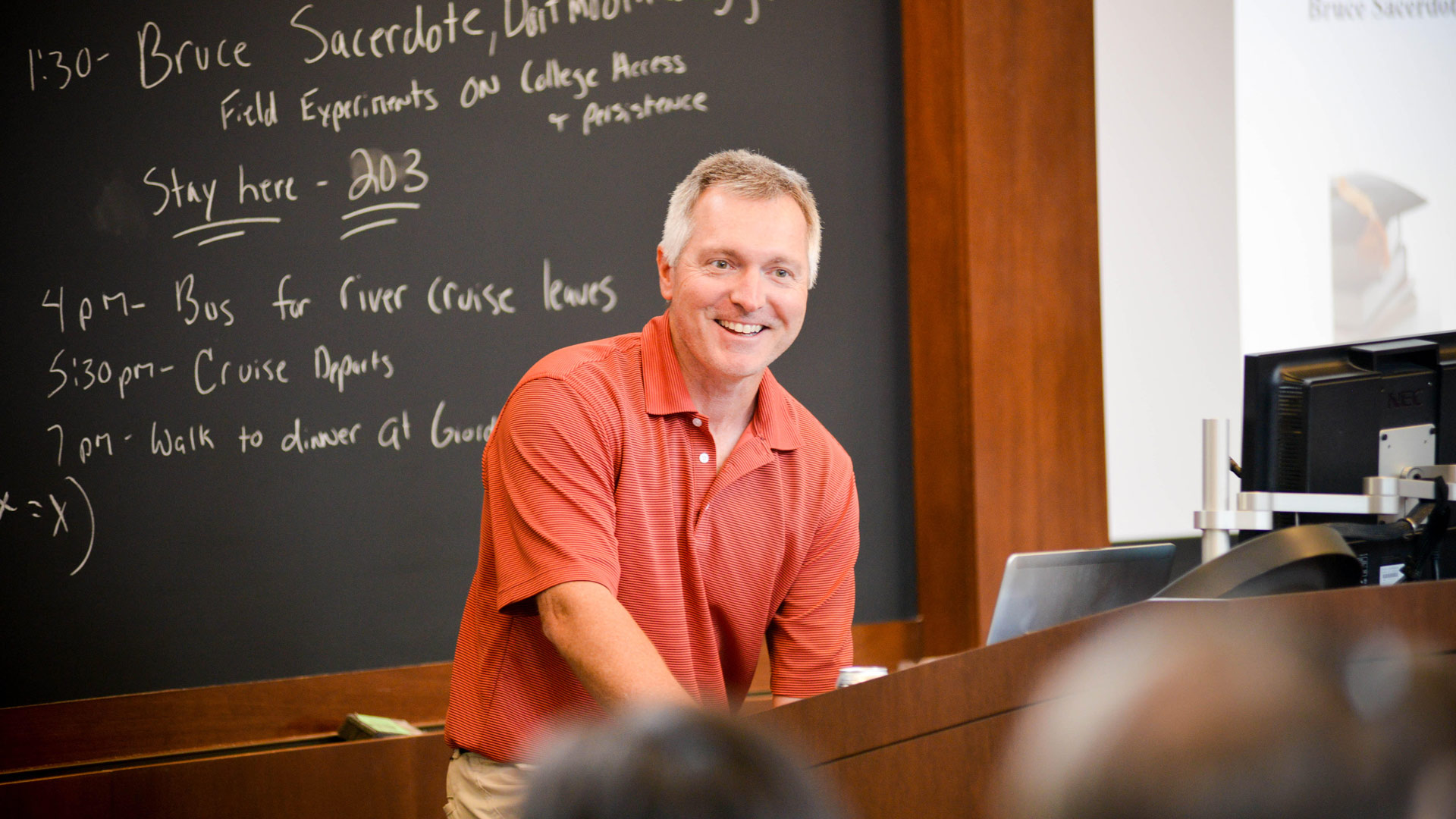 Man in orange shirt smiles at audience member as he gives lecture.