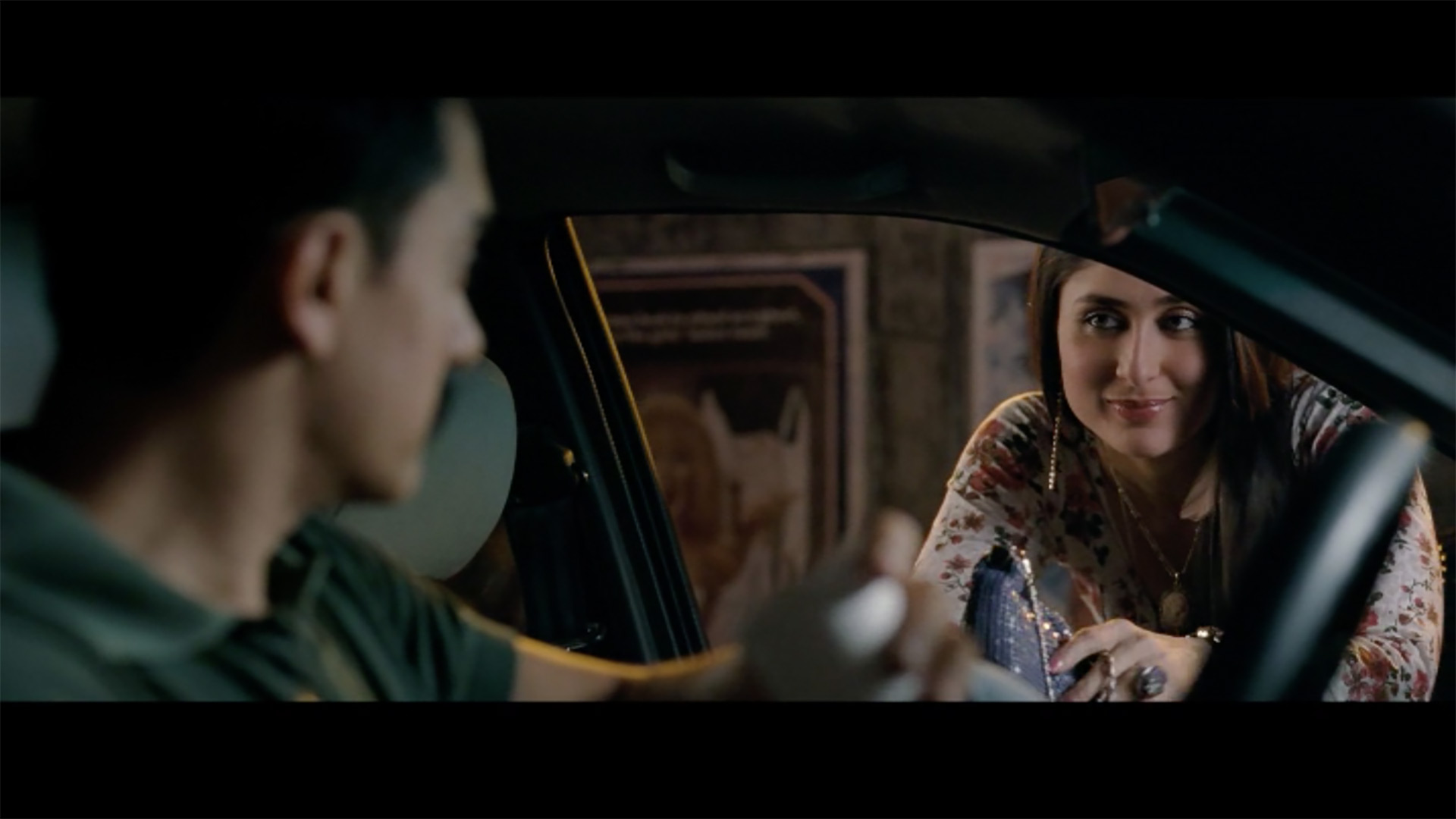 A woman with a slight smile looks into the driver side window of a car at a male driver at night