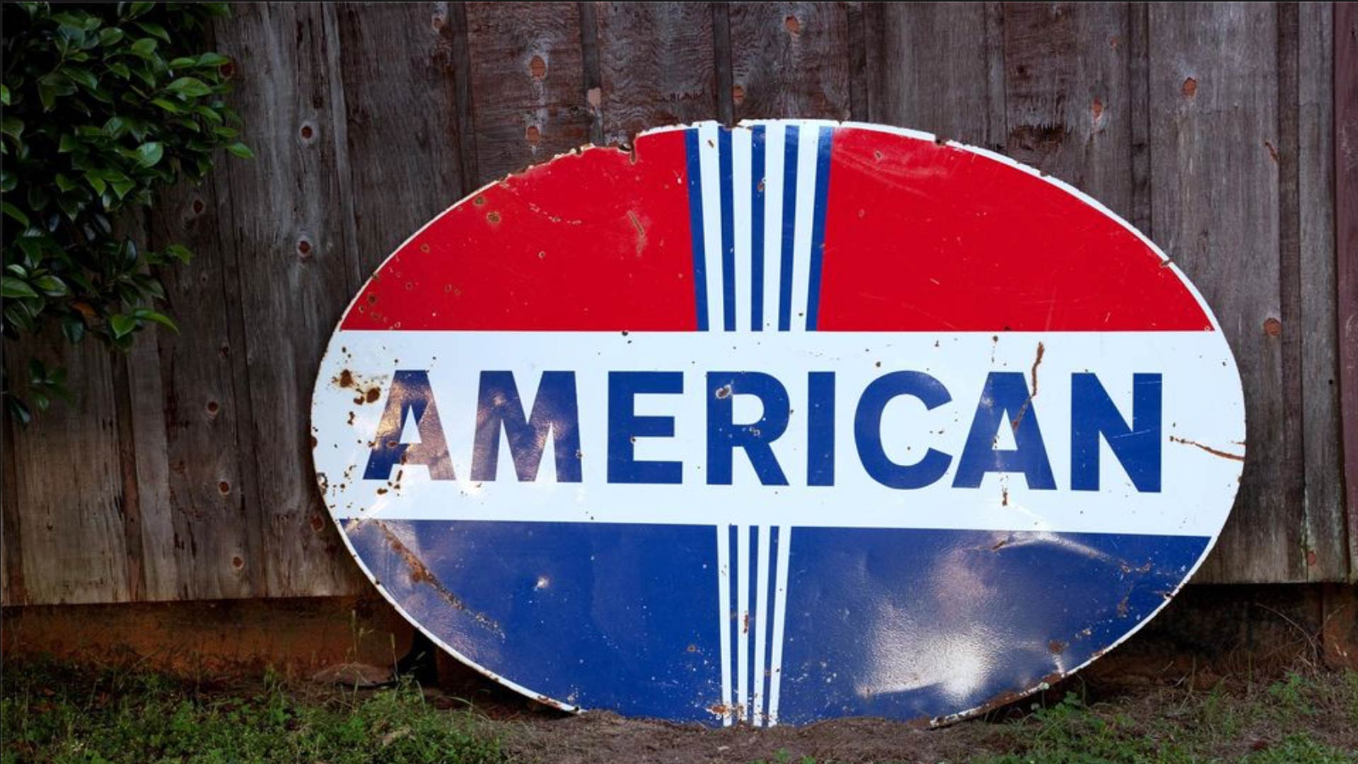 a rusted and dented oval sign with AMERICAN written on it leaning against a fence