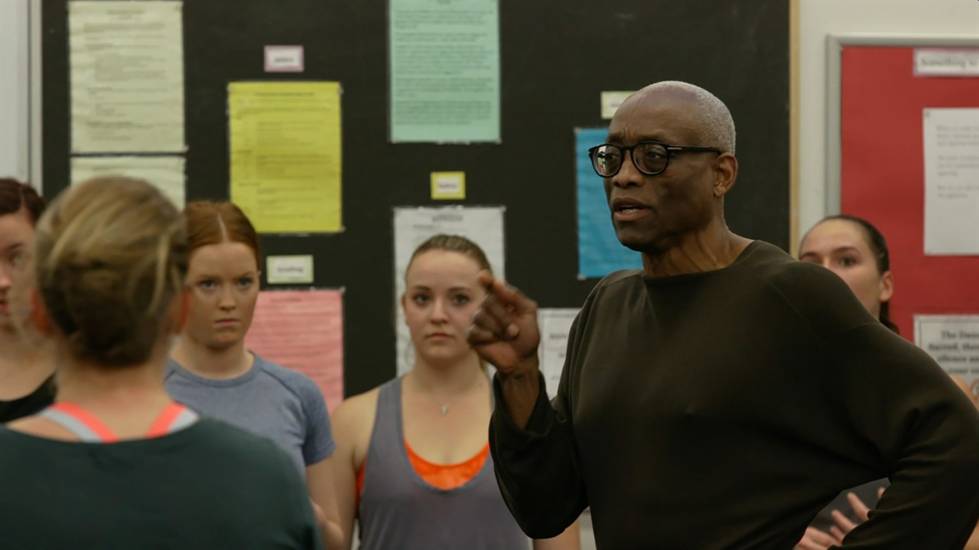 Dance instructor speaks to students who stand around him in a classroom