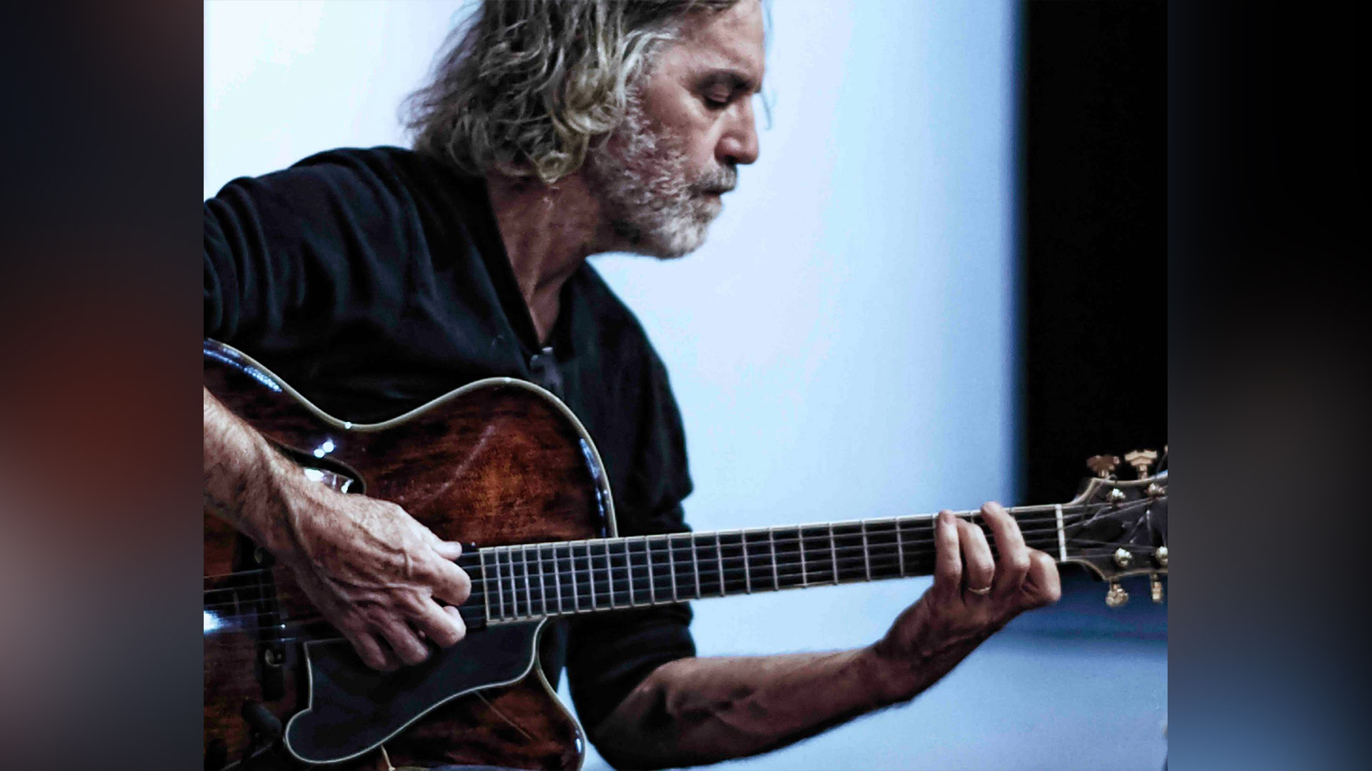 man with long hair plays a guitar in front of a light wall