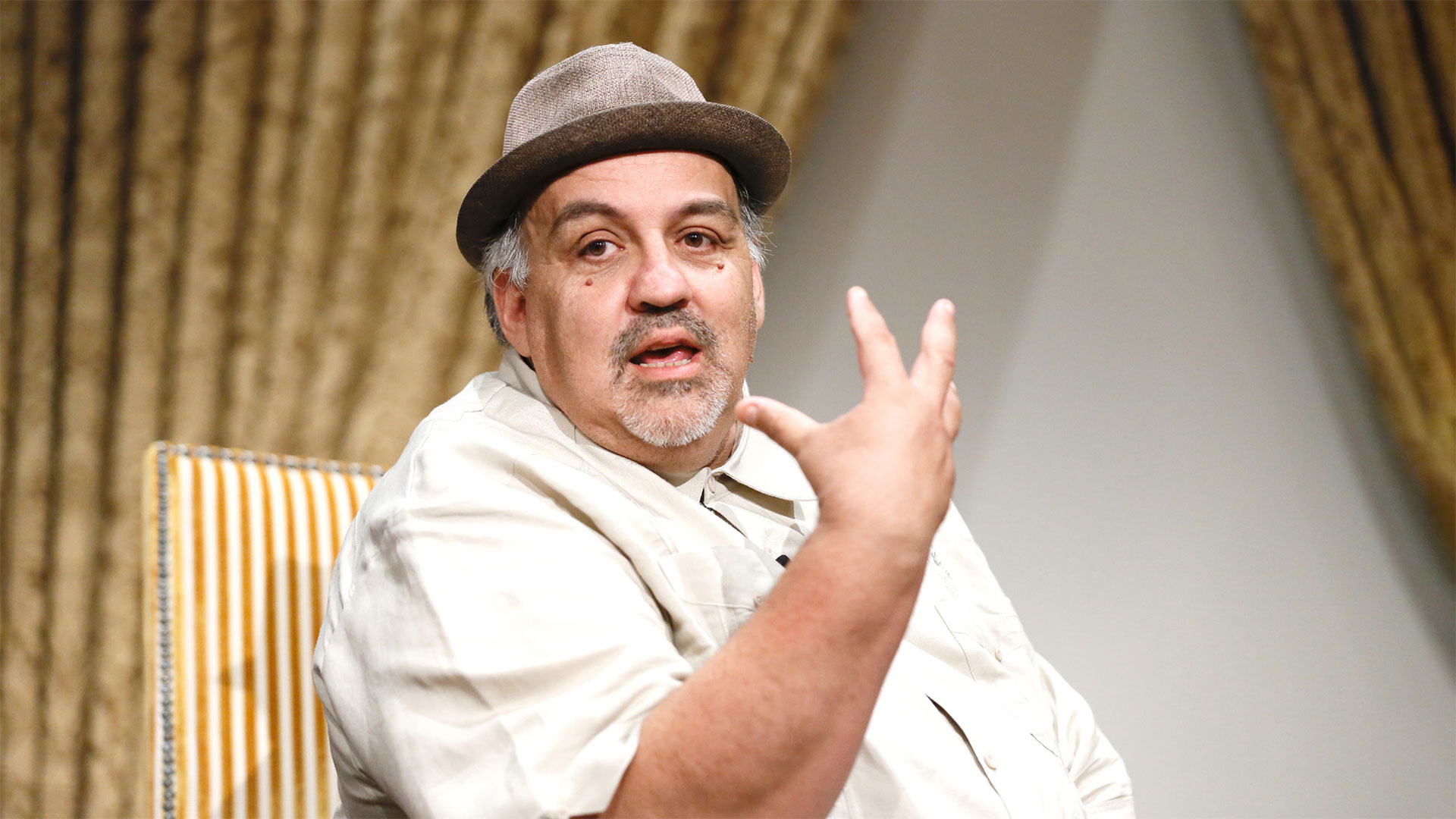 Man sitting in a chair gesturing and speaking in front of a curtain