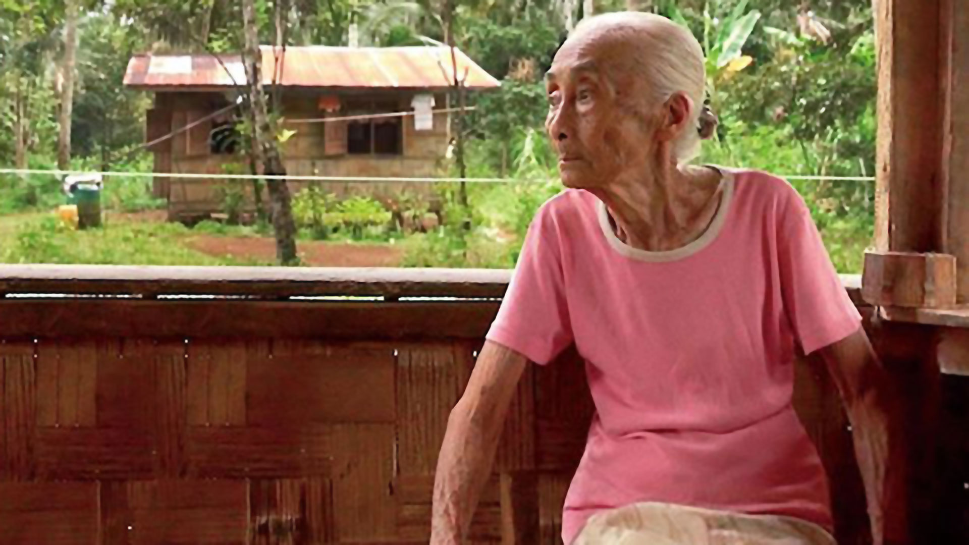 an old woman sitting on a porch, looking to the left, with a tropical forest and a small house in the background