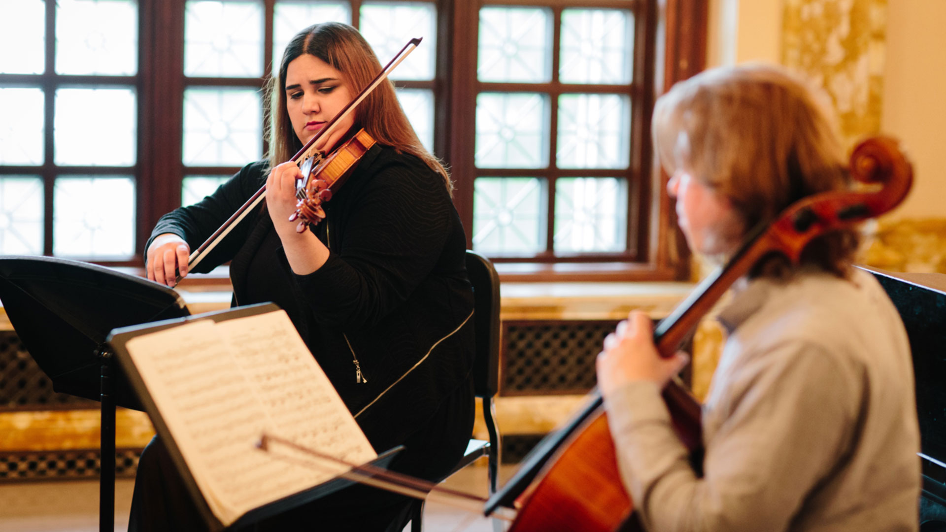 Two women play stringed instruments with music stands in an indoor venue