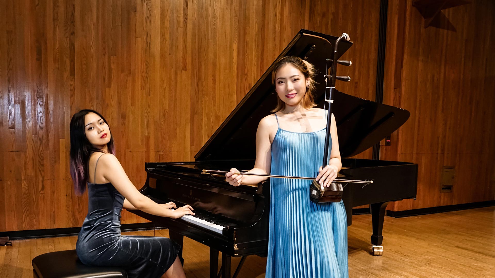 Two women of Asian descent dressed formally on a stage with wooden walls, one at a grand piano, and one holding an erhu
