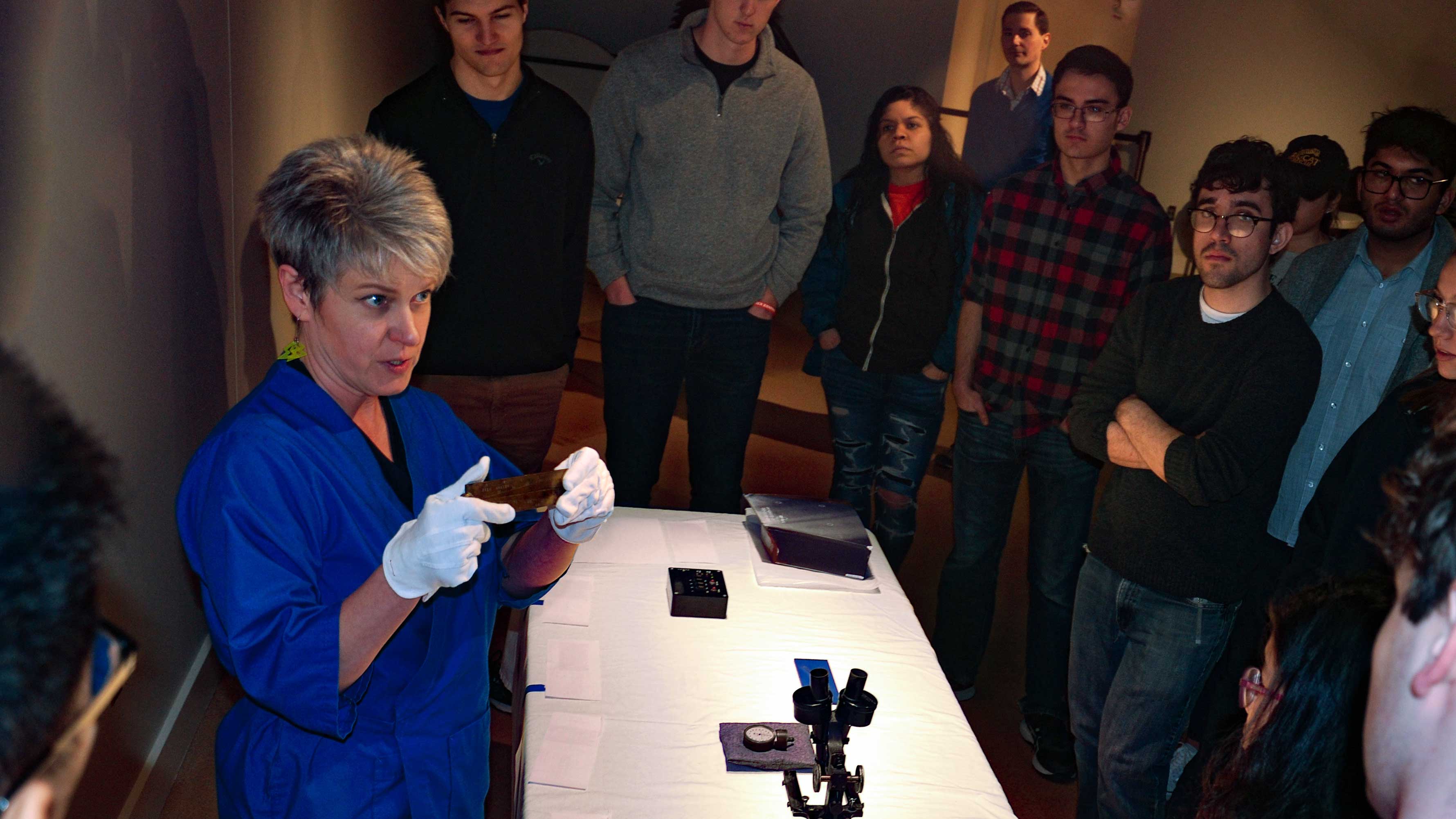 museum staff speaks to students gathered around a table with a microscope