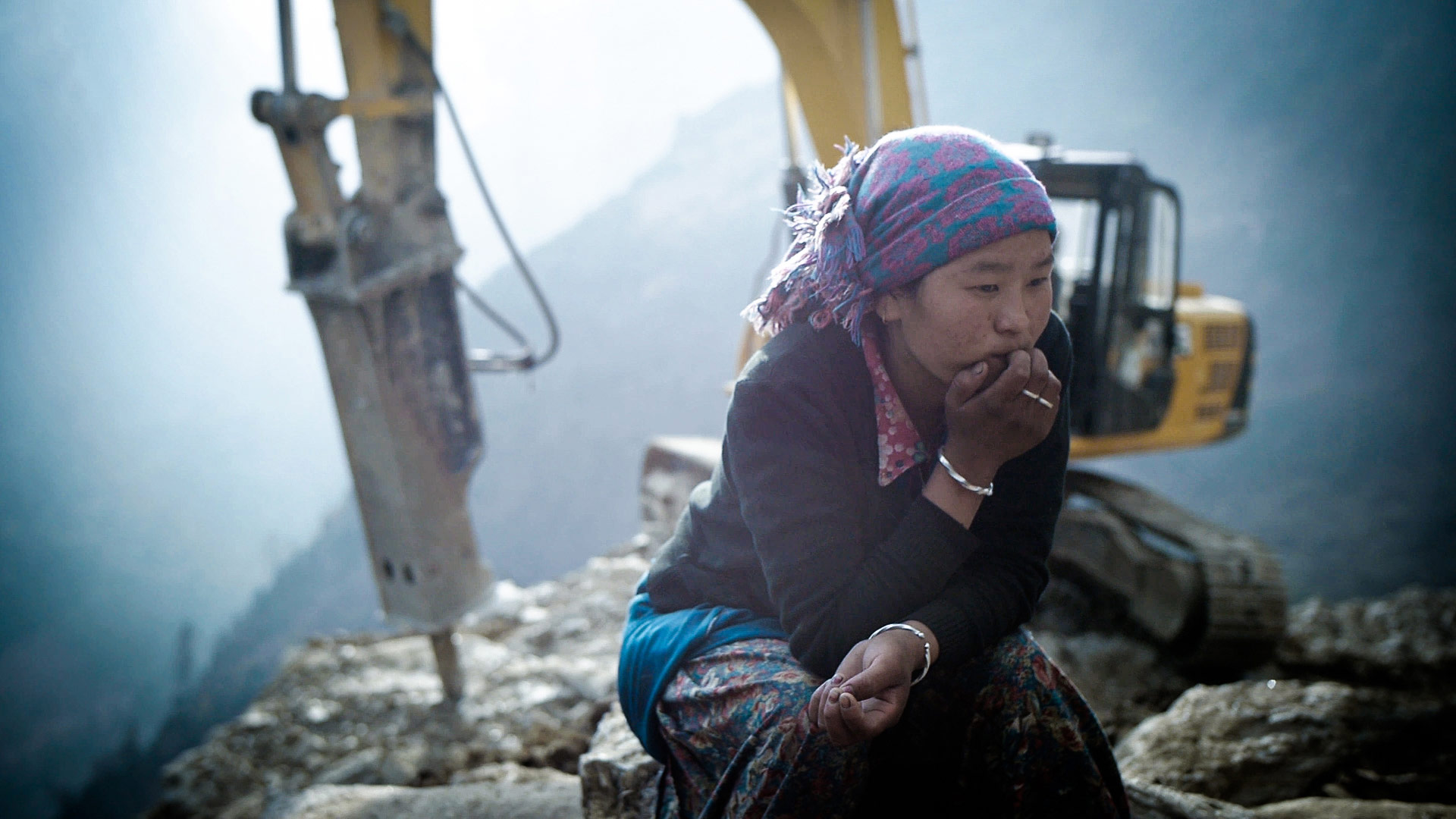 A woman crouches in thought in a rubble pile with an excavating machine behind her