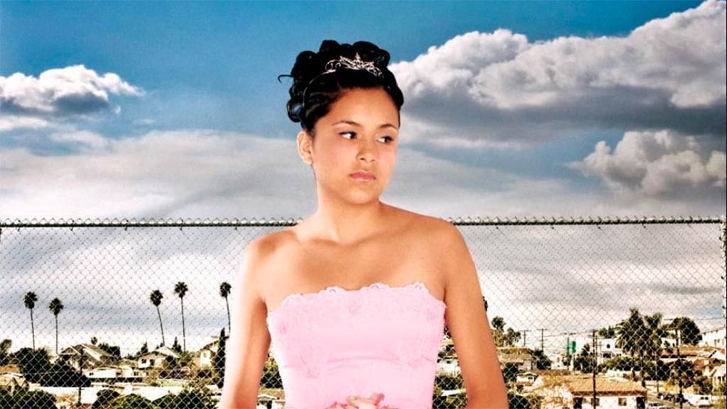young woman with a pink formal dress in front of a chainlink fence and vista of residental buildings and palm trees