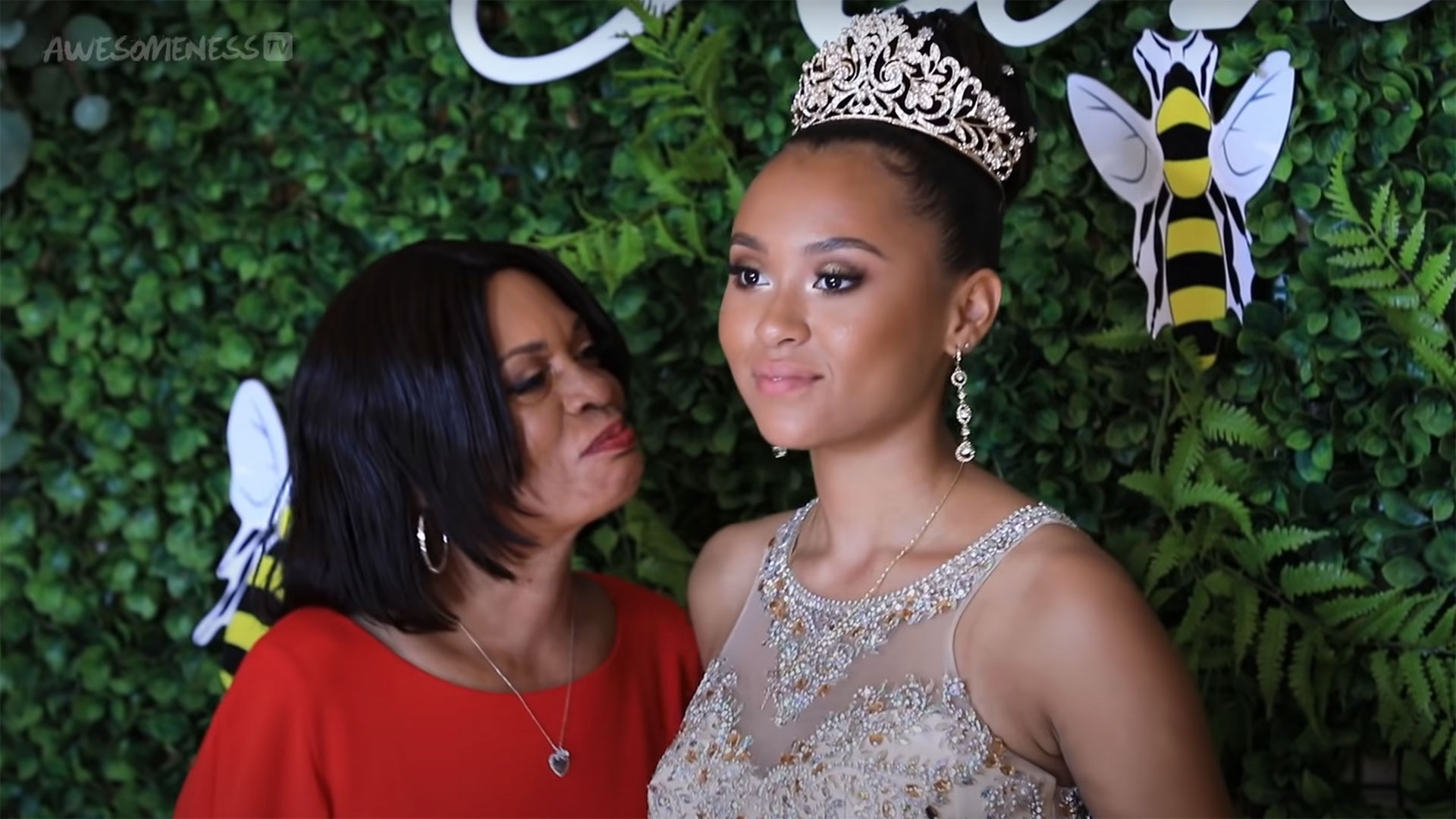 young woman with tiara poses in front of a greenery wall while older woman, potentially mother, looks on lovingly