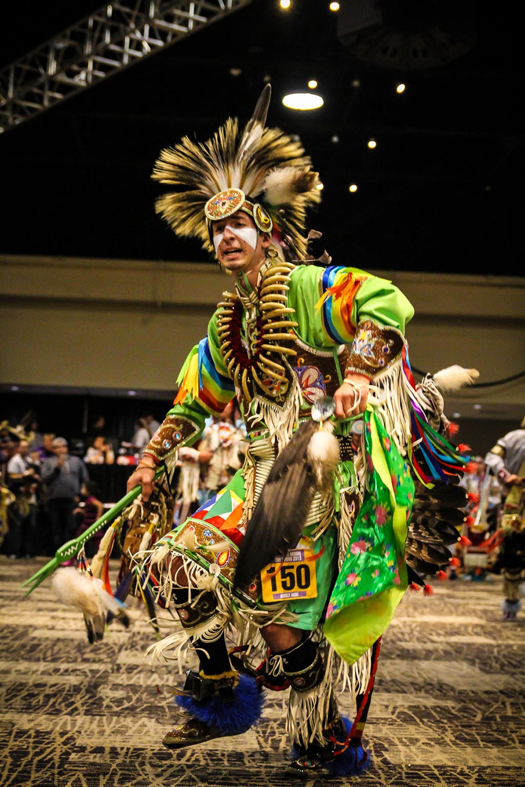A man wearing bright green traditional powwow clothing while dancing in a conference room.