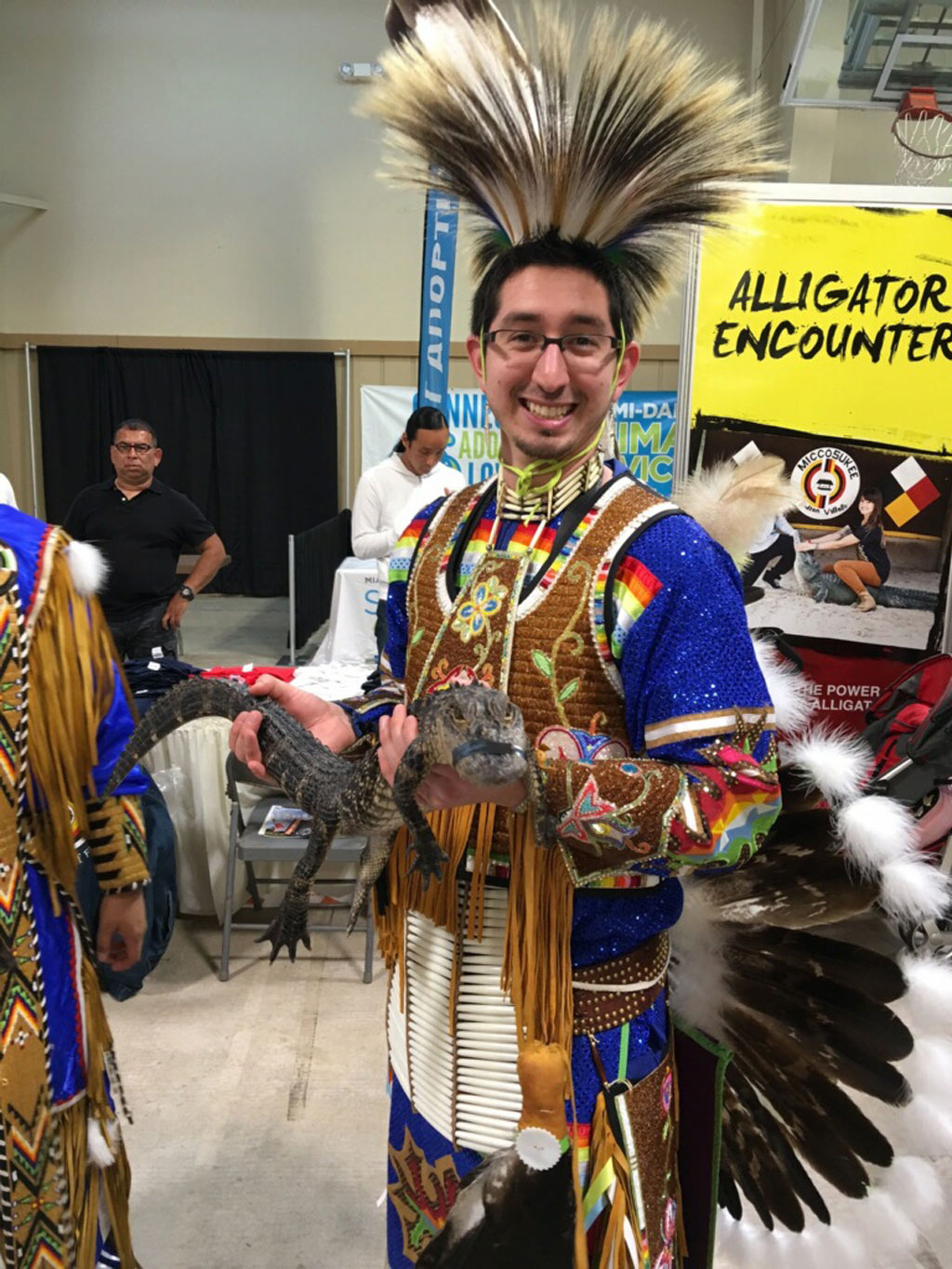 A man with glasses wearing colorful traditional powwow clothing while holding a baby alligator 