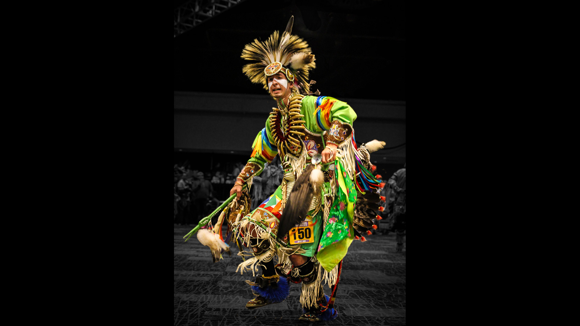 A man wearing bright green traditional powwow clothing while dancing in a conference room.