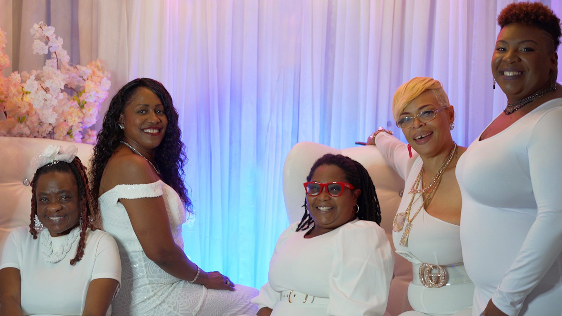 Five African American women in white dresses sitting in front of a pastel backdrop.