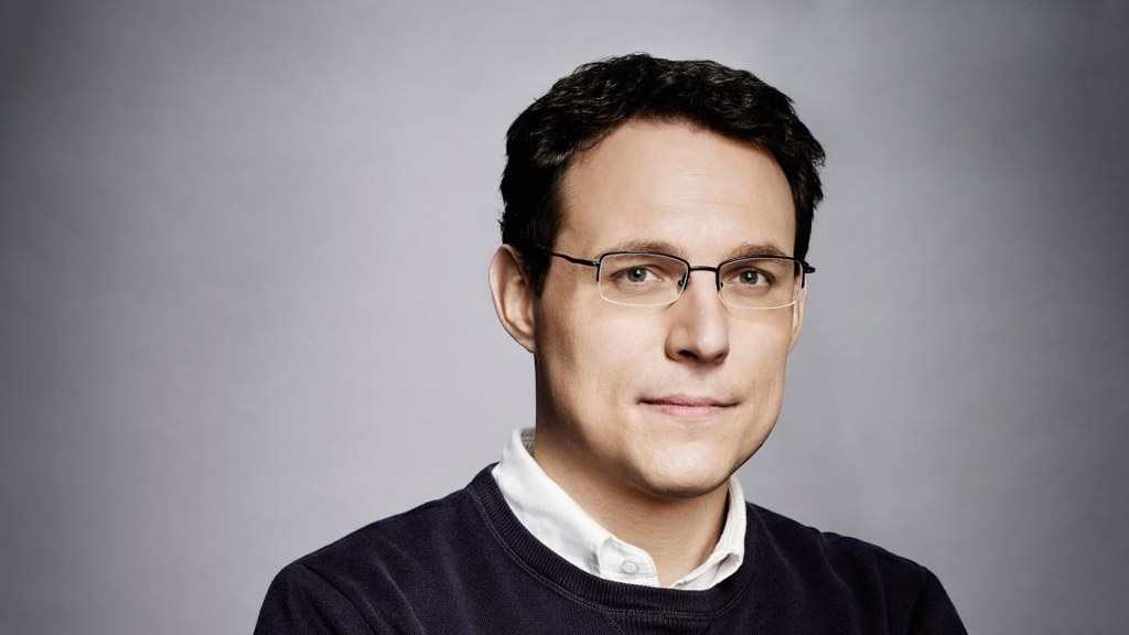 headshot of a man with a neutral expression, glasses, and a sweater in front of a gray background