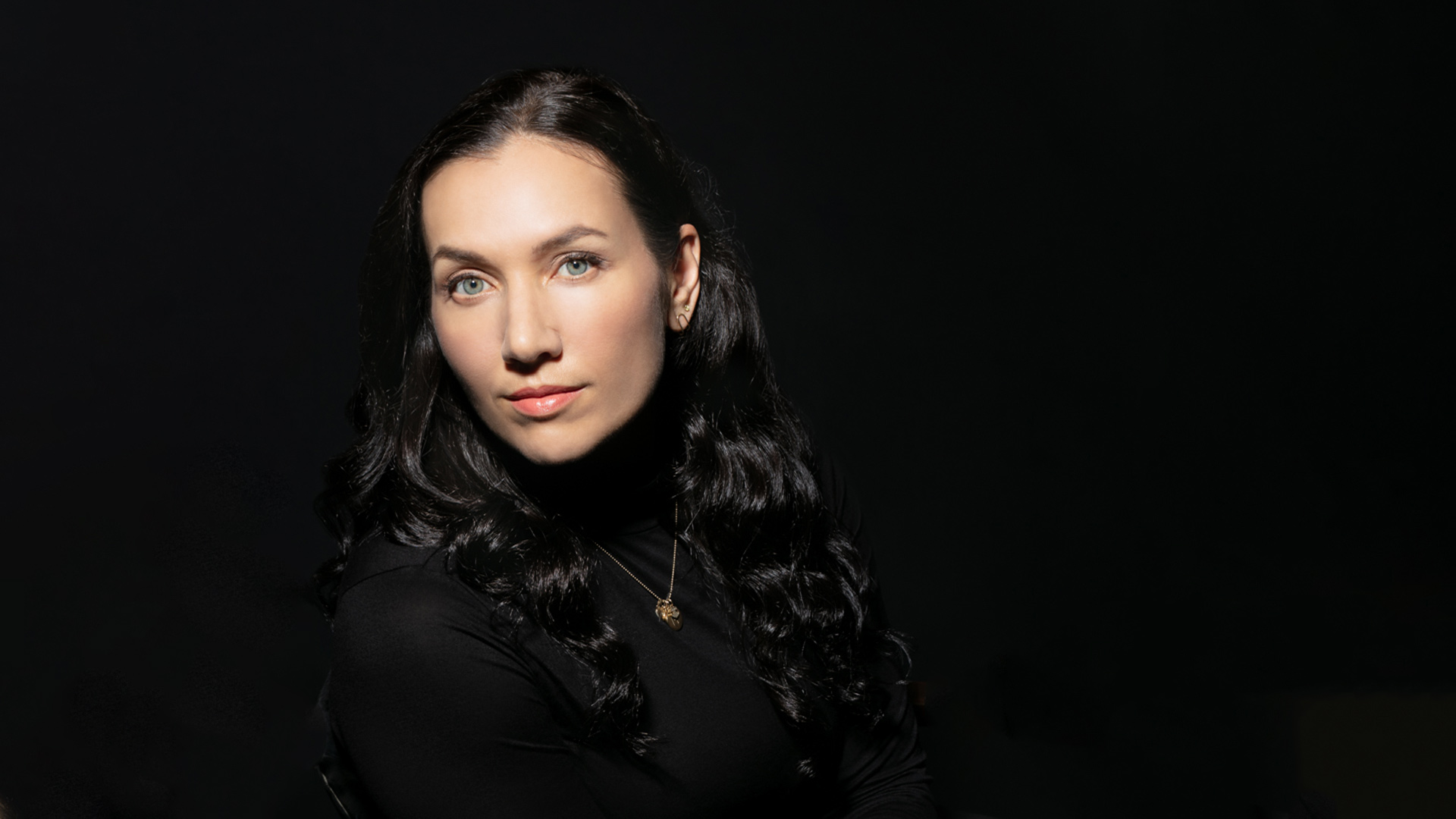 woman wearing a black turtleneck and gold necklace sitting in front of a plain black background