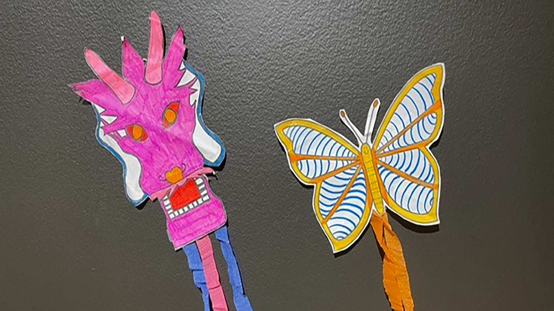 pink paper kite in the shape of a dragon face with blue and pink streamers, next to an orange butterfly kite with orange streamers