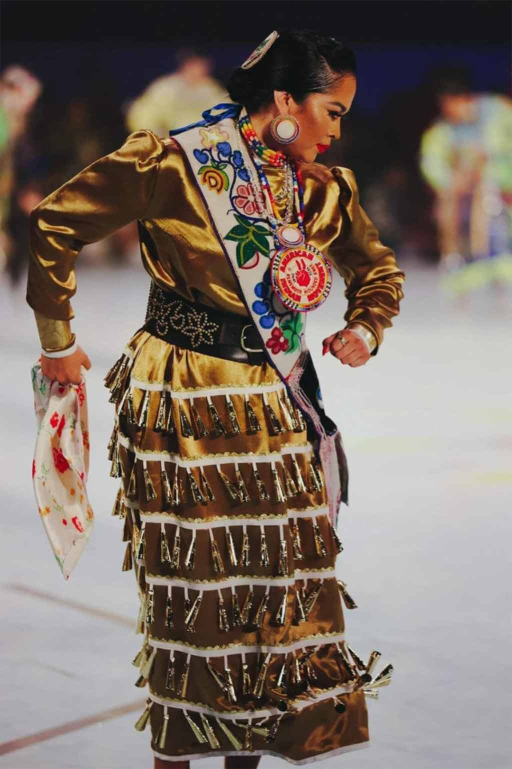 woman with beaded sash dancing in a gold dress with rows of golden noisemakers
