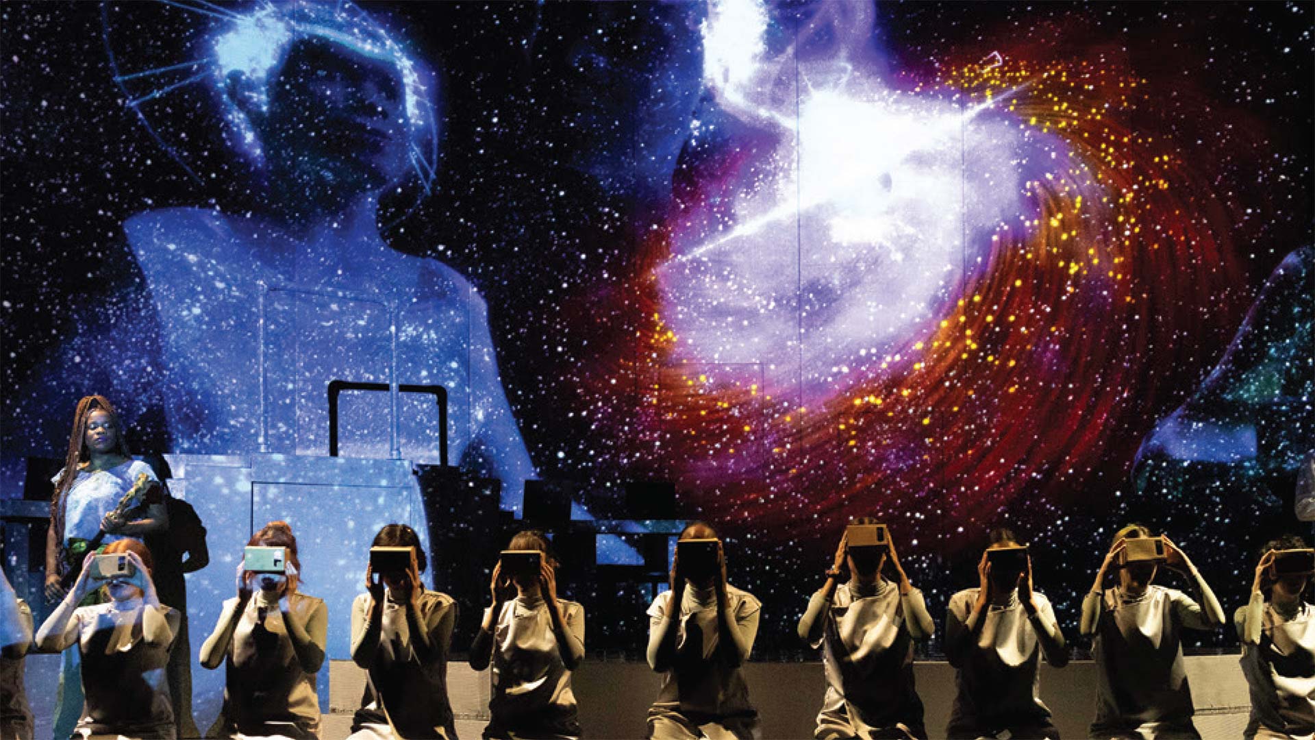 stage performance with starfield visualization and 9 women kneeling and looking into viewboxes alongside a guitarist