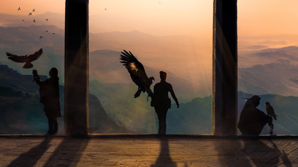 silhouettes of three people holding a falcon with mountains and sunshine behind them