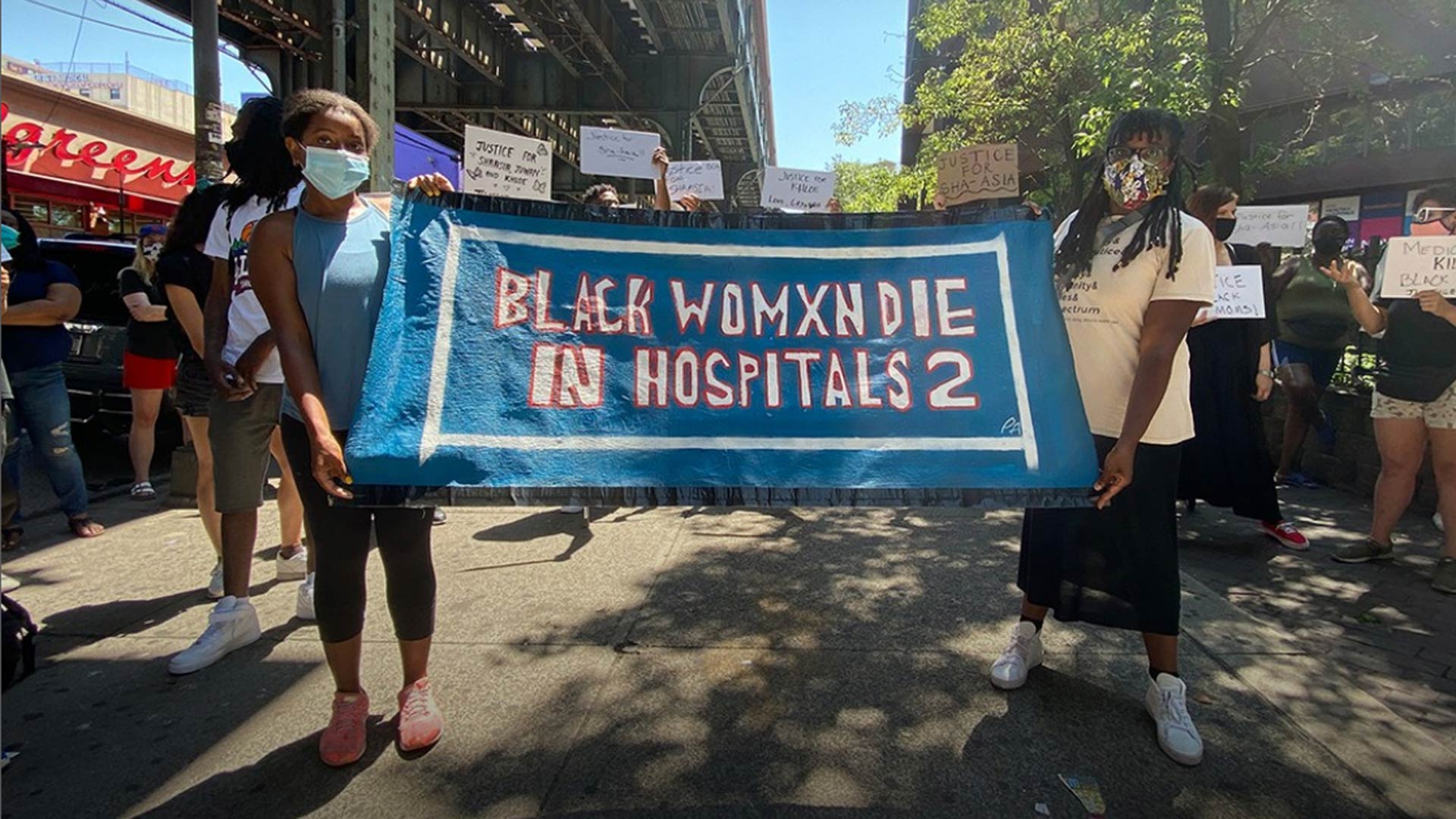 two black women holding up a banner that reads "black womxn die in hospitals 2"