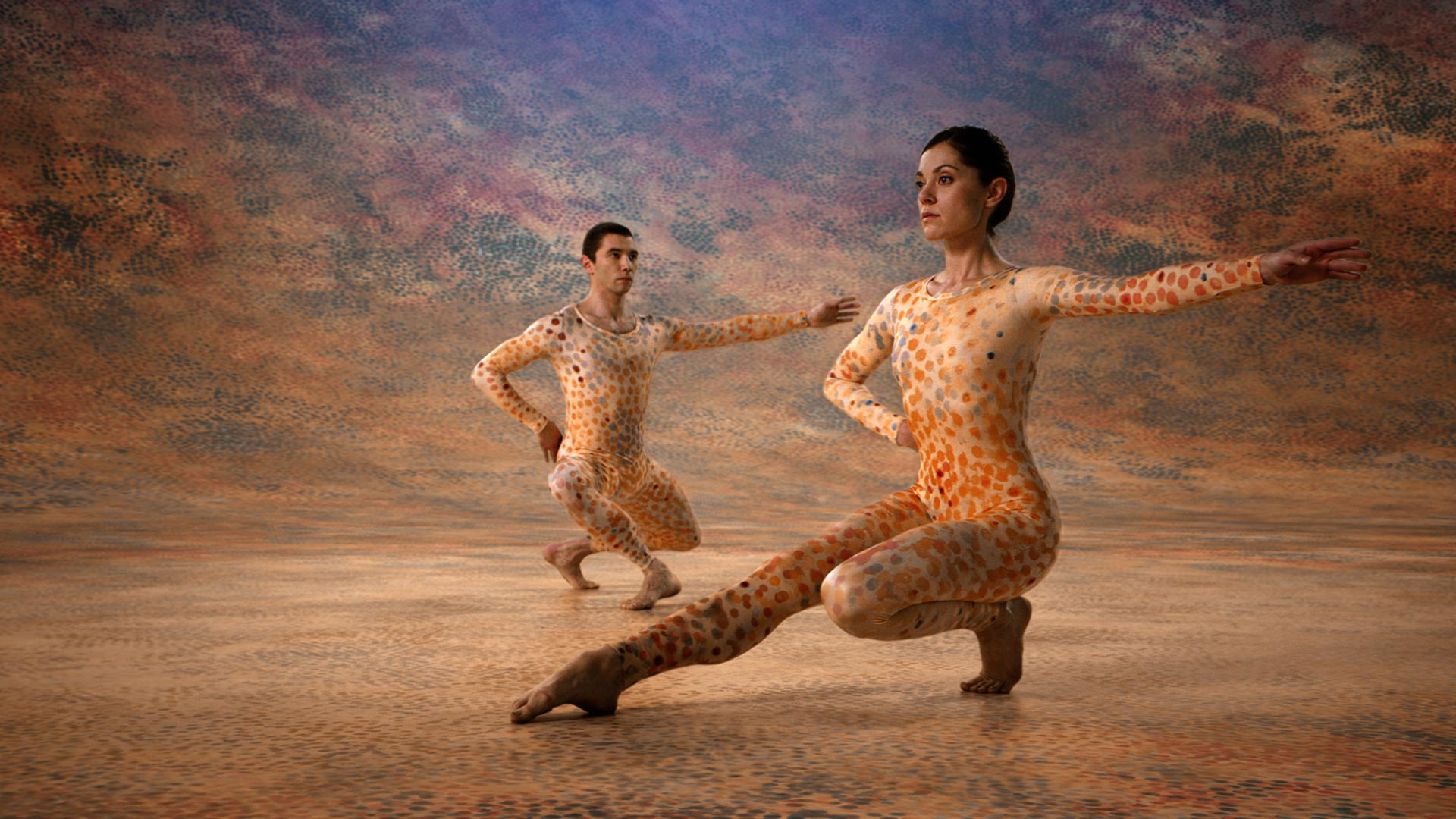 Two dancers in tan, dotted body suits crouched on floor with one arm extended