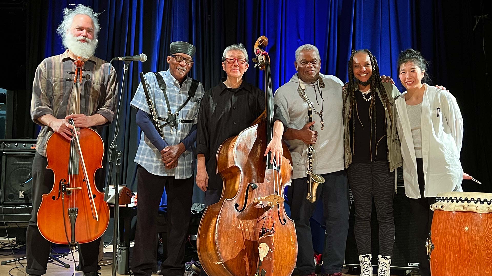 Six members of the MIYUMI Project ensemble pose on stage for a picture with stringed and reed instruments as well as a drum