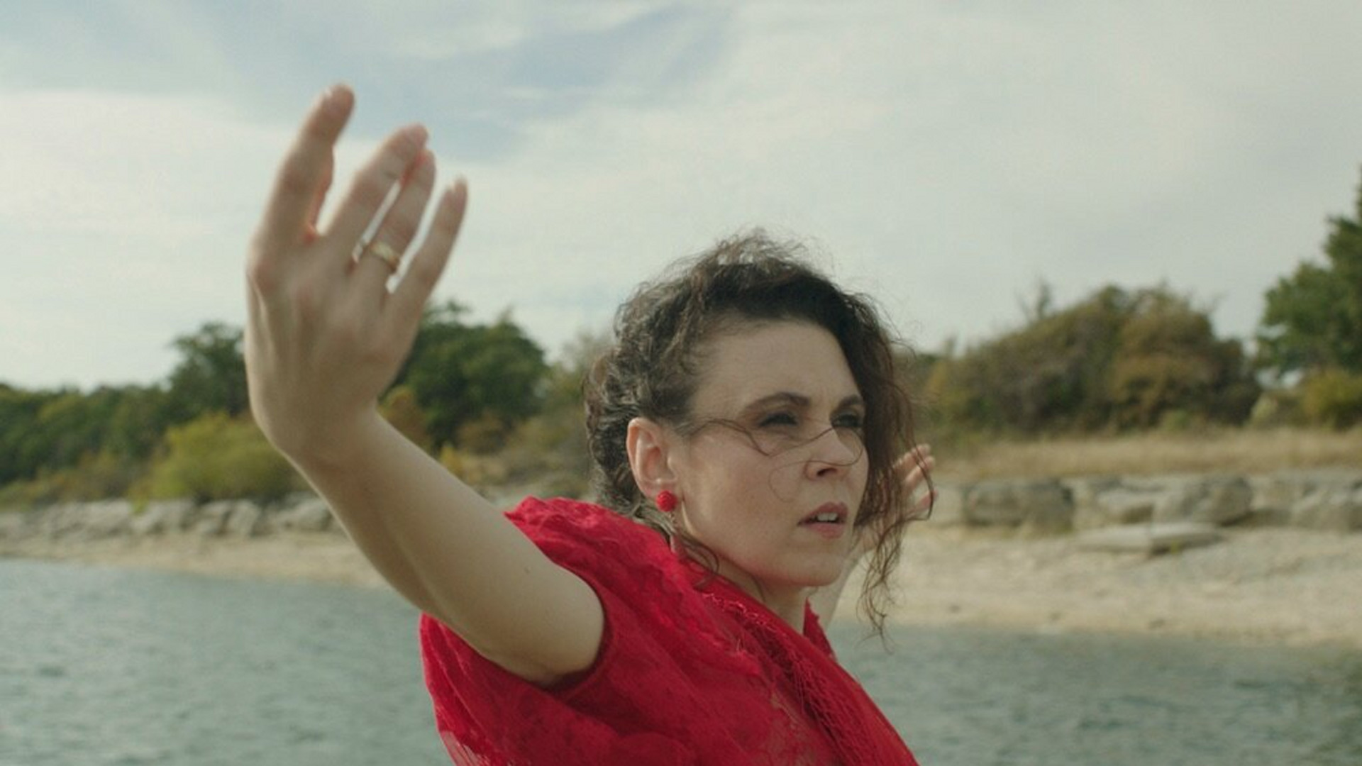 Woman wearing a red blouse and red earrings with outstreched arms in front of a beach