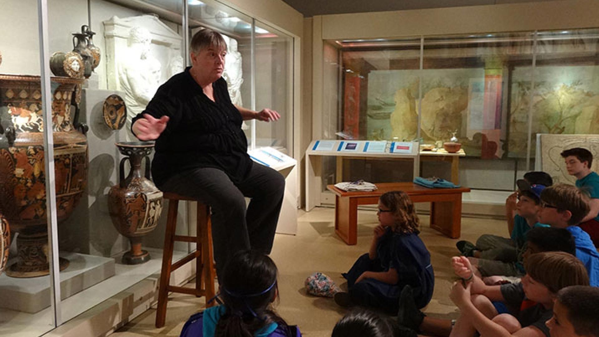 woman sitting on a stool in front of a group of children with a case of artifacts behind her