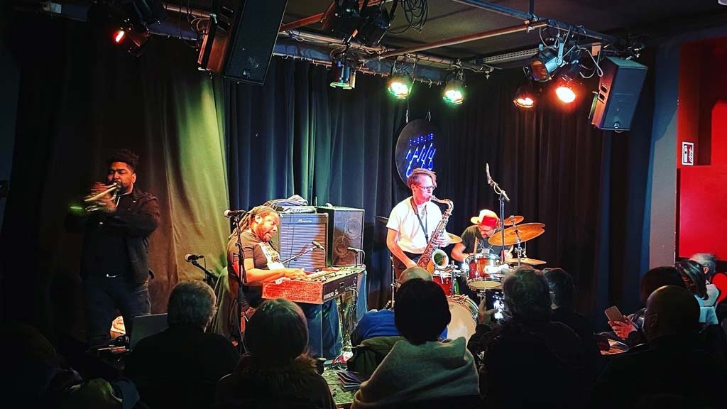 four musicians on a stage with audience members in the foreground and colorful lights