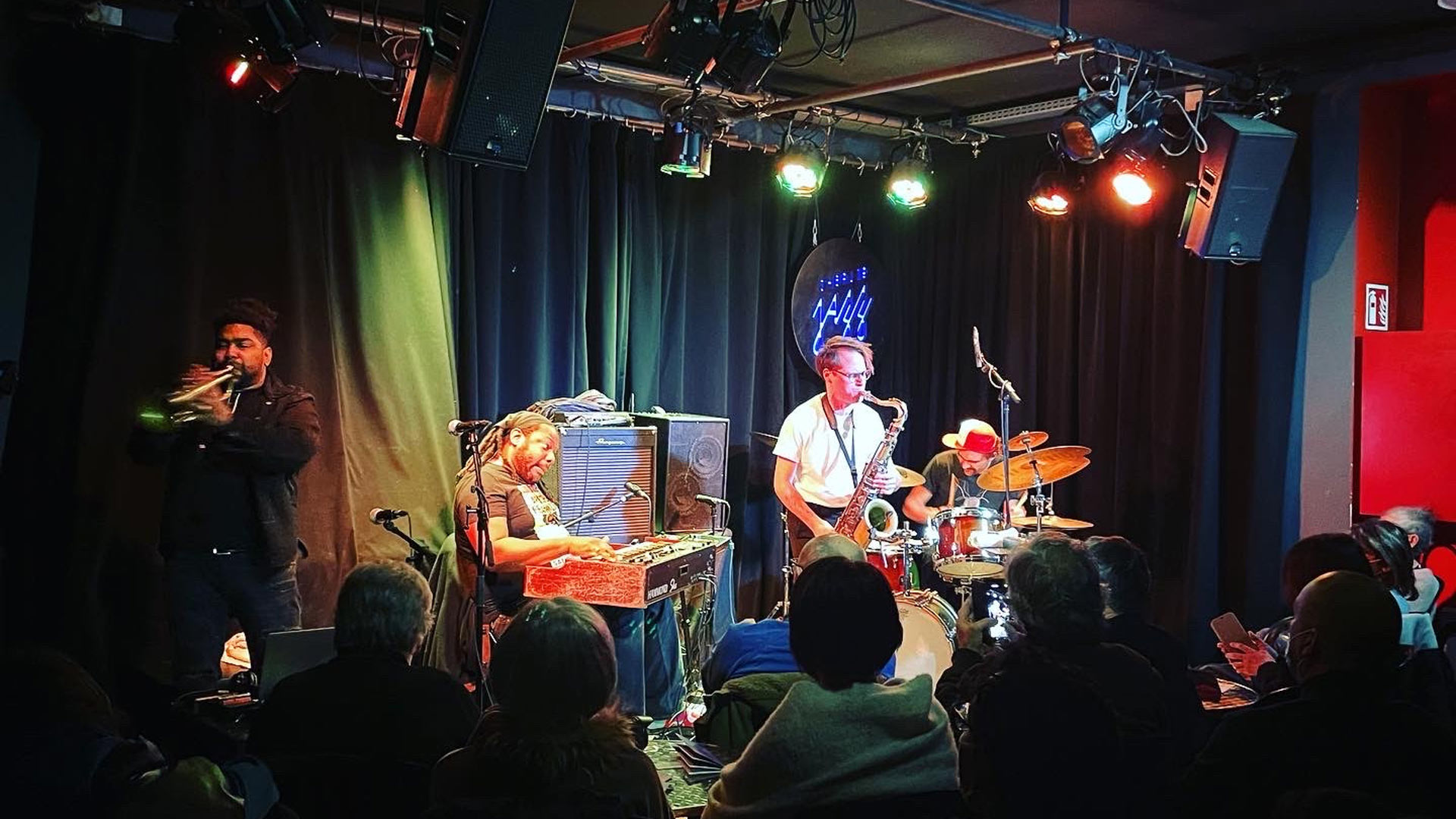 four musicians on a stage with audience members in the foreground and colorful lights