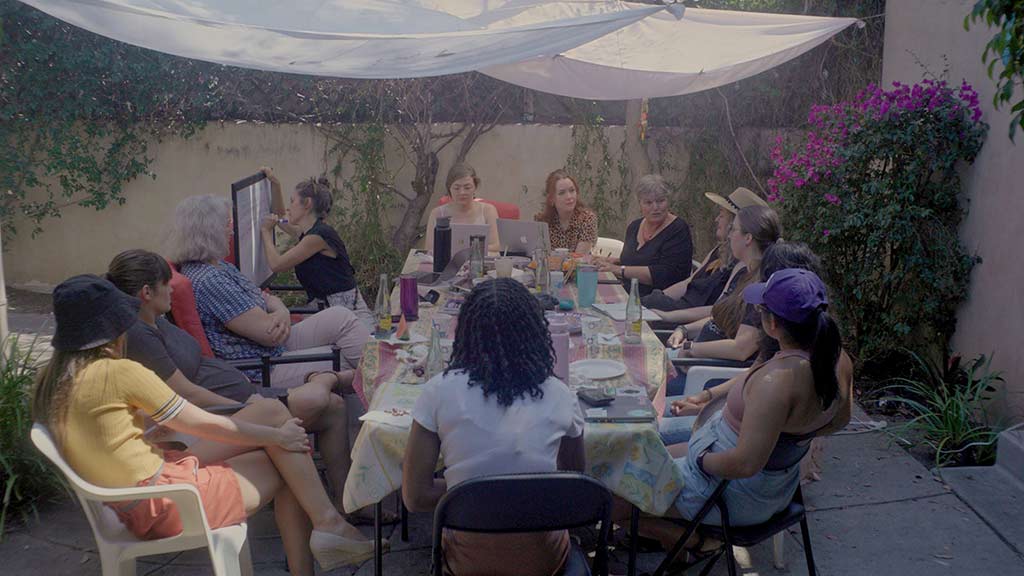 A group of women sit around a table discussing ideas while one writes on a whiteboard