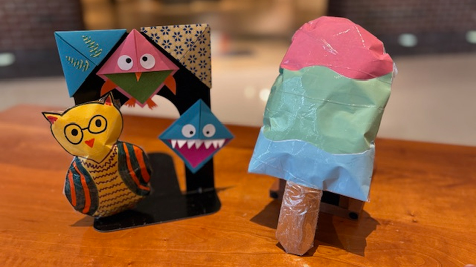 Triangle patterned paper bookmarks in the shape of animals on display next to a pink, green, and blue popsicle on a brown table
