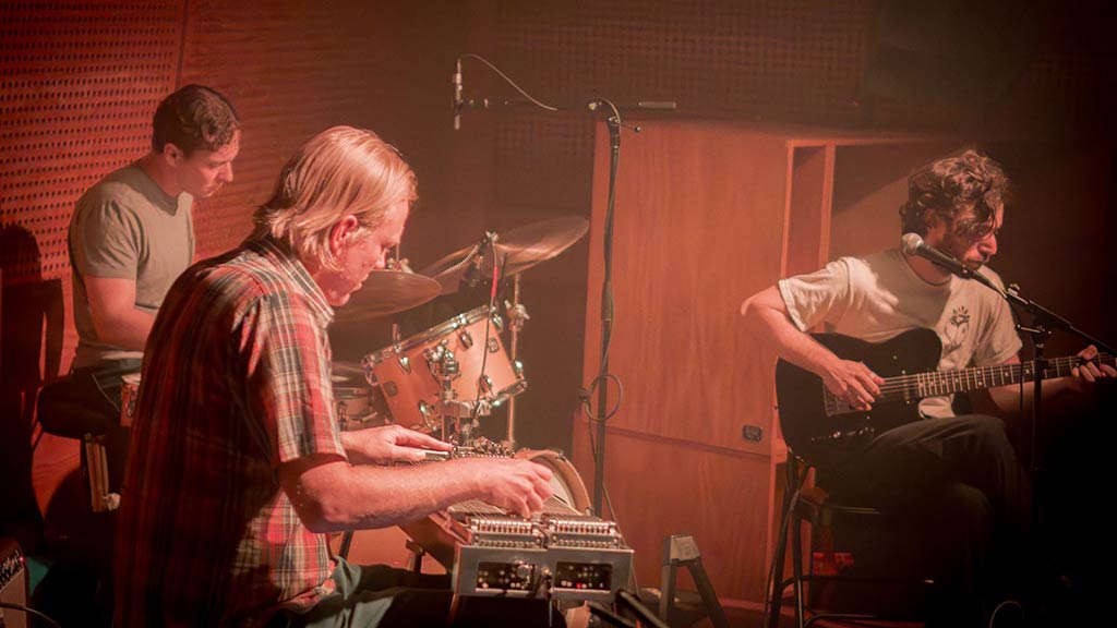 three male band members in casual clothes play drums, keyboard, and guitar in a hazy studio setting