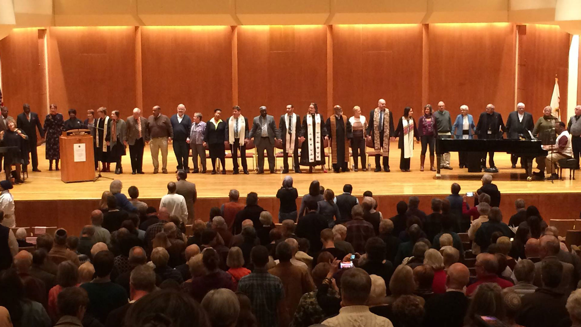 clergies from different religions on stage lined up while holding hands.