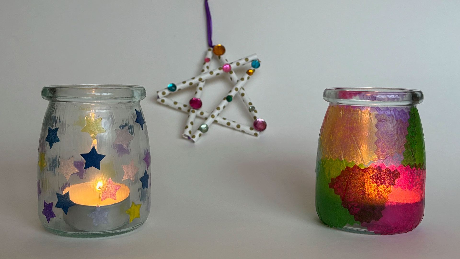 a paper straw star craft hangs behind jars decorated with stars and translucent colored paper illuminated via tea candles