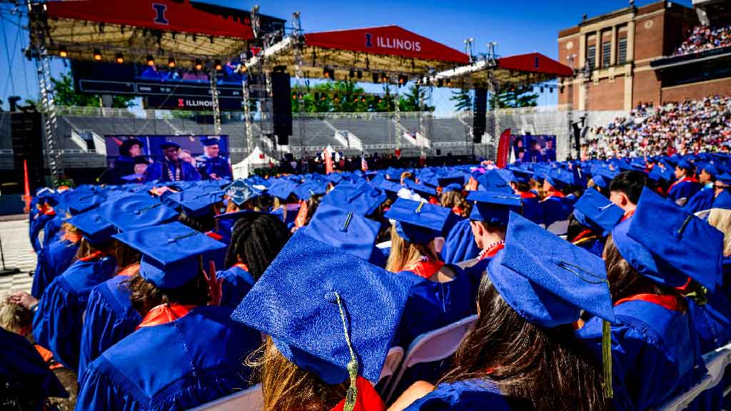 a view of an outdoor graduation ceremony at memorial stadium with a sea of blue graduation caps in the foreground
