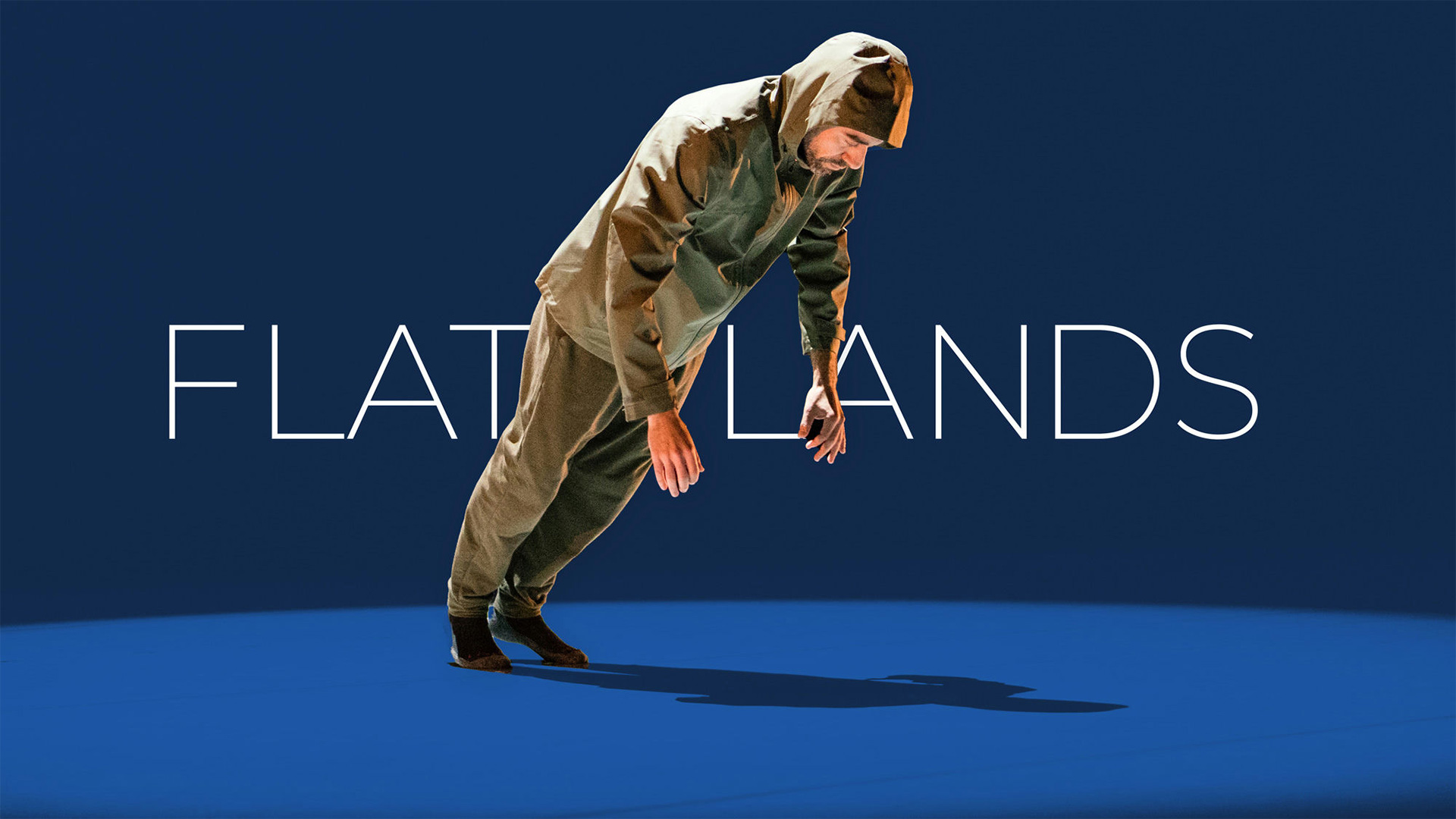 dancer wearing a hooded jacket and pants leaning forward over a blue spotlight in front of FLATLANDS text
