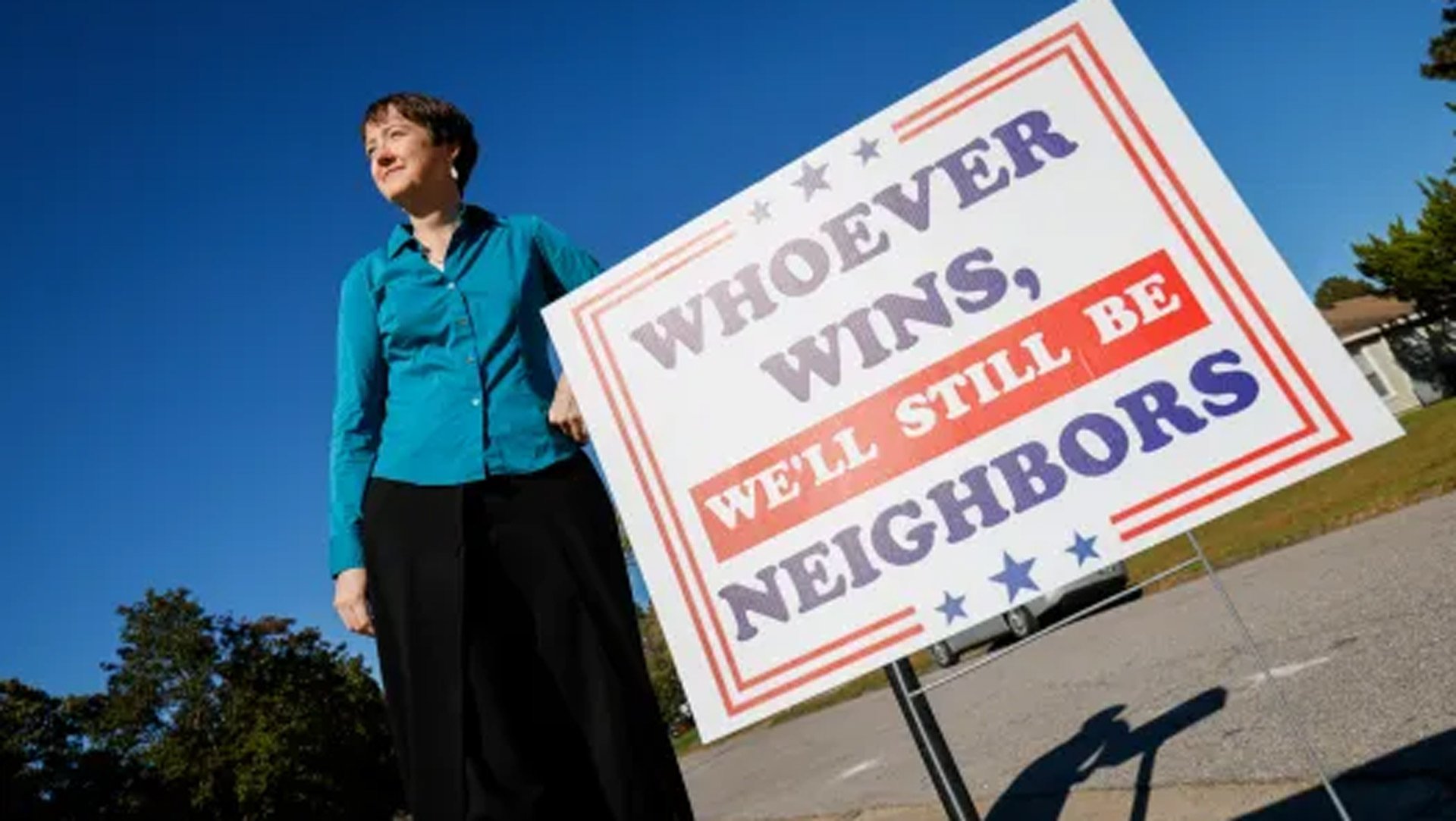 A woman in a blue shirt looking into the sun while standing next to a red, white, and blue sign.