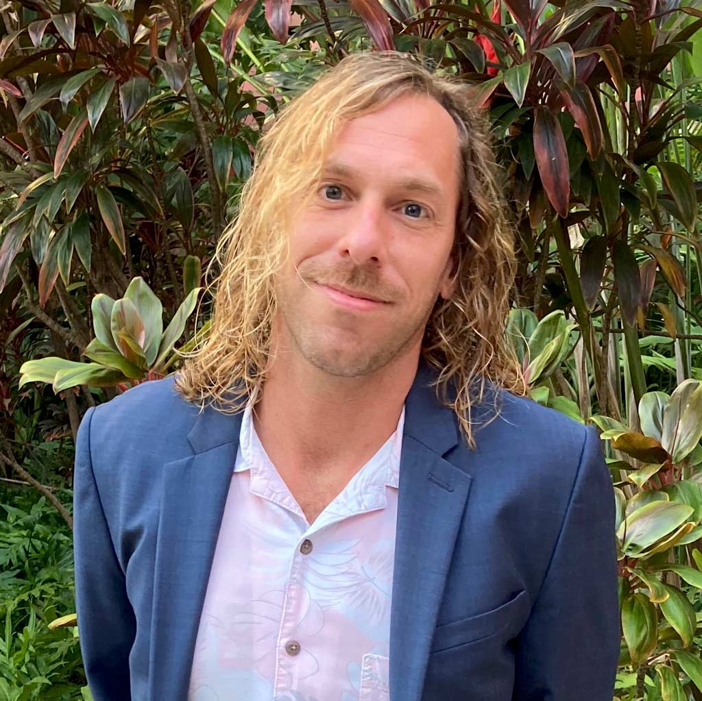 a head shot of a man with long blonde hair in a blue blazer looking straight with a smile.