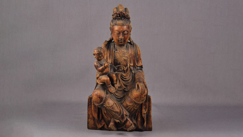 a wooden carving of a Buddhist deity sitting and dressed in a robe and various accessories while holding a child.
